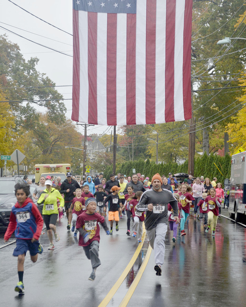 Entrants in the Fanwood Family Fun Run, part of the borough's 5K race activities, stream under the Fanwood Fire Dept.'s large American flag on LaGrande Avenue in Fanwood, NJ, Sunday, Oct. 25, 2015. (Photo by Brian Horton)